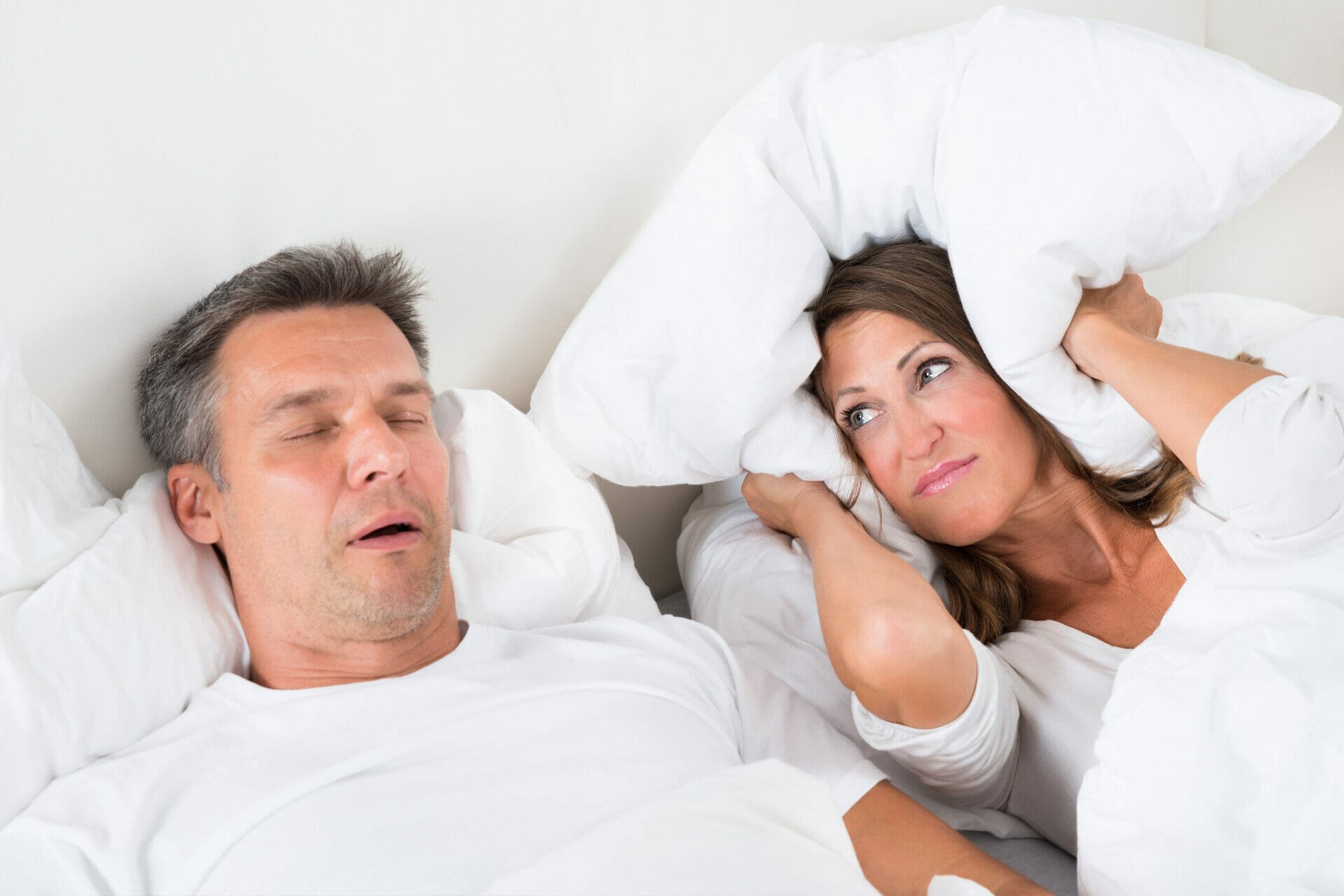 A couple lying in bed. The man is snoring whilst sleeping and the woman is covering her ears with a pillow in frustration